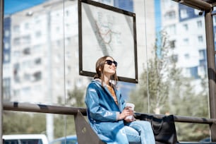 Junge stilvolle Frau, die auf die öffentlichen Verkehrsmittel wartet, während sie an der modernen Straßenbahnhaltestelle im Freien sitzt