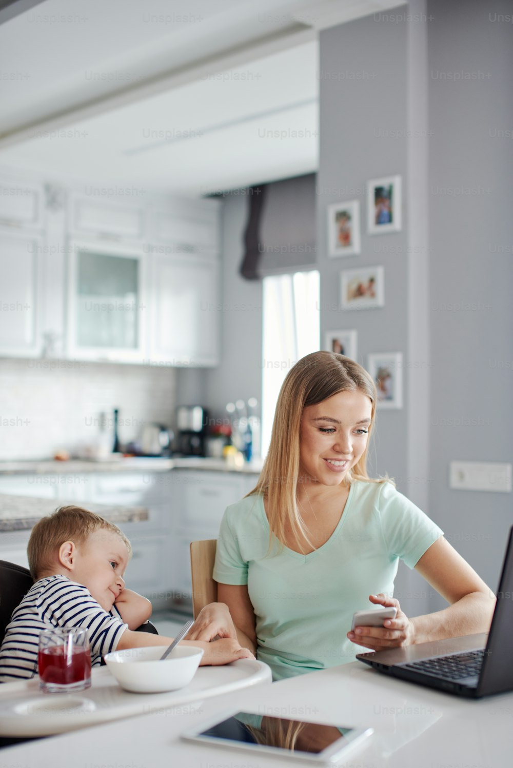 Portrait of mid age caucasian blond woman with child being busy with phone and computer e and feeding the baby. They are at home, wearing pyjamas