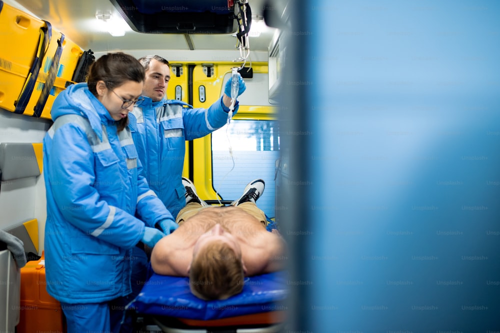 Una de las dos jóvenes paramédicas que examinan a un hombre inconsciente sin camisa en camilla mientras su colega sostiene el gotero