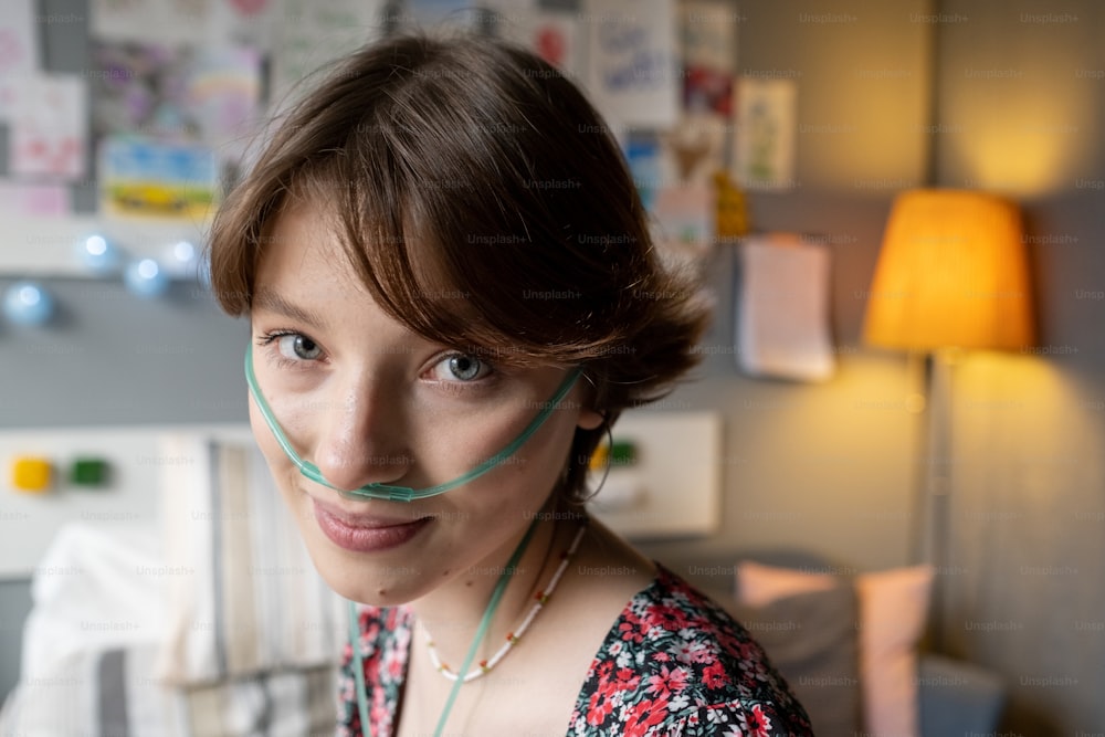 Cara de una adolescente sonriente con un tubo de oxígeno en la nariz mirando a la cámara contra el interior de la sala del hospital