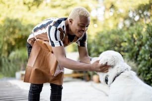 Black man playing with a dog. Concept of gardening and leisure. Young smiling handsome guy wearing apron. Idea of modern domestic lifestyle on summer sunny daytime.