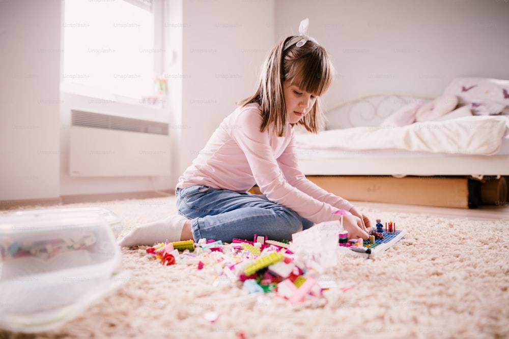 Beautiful thoughtful little toddler girl sitting on the carpet of her bright room and playing with plastic toys.
