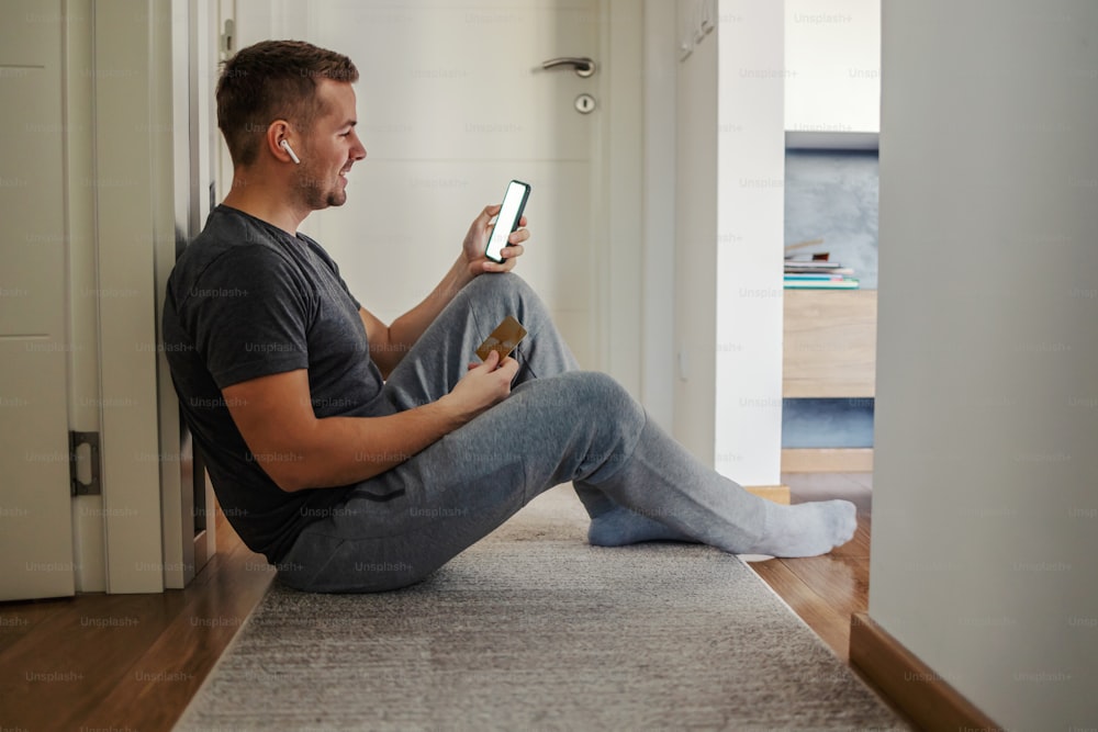 Online account verification. A casual man in a modern home sits on the floor of entry hallway and holds a card and a phone in his hands. Online checking status