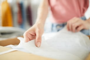 Mãos de jovens designers precisos dobrando o papel do pacote enquanto embrulha roupas recém-feitas antes de enviá-las ao cliente