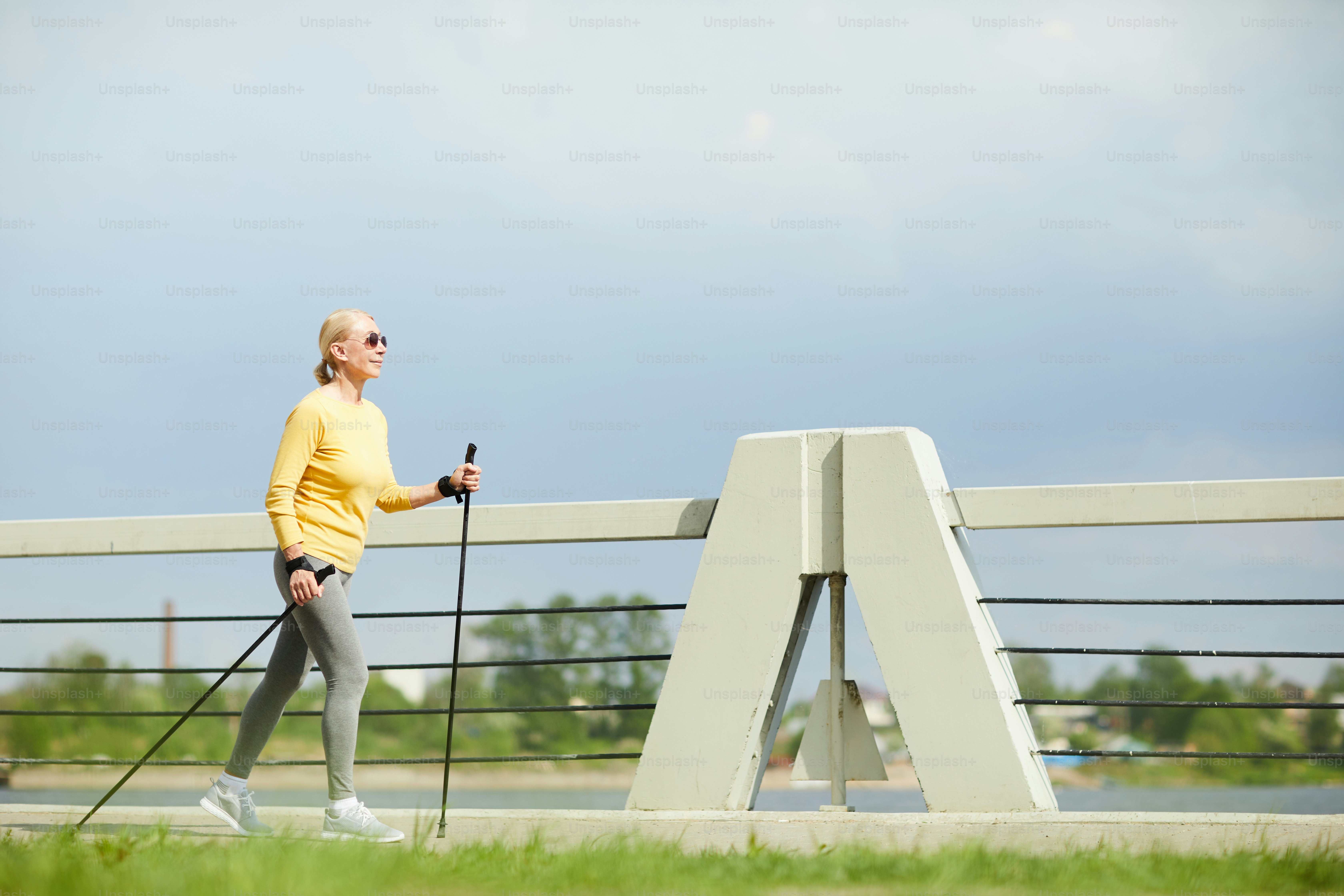 AGed sportswoman in pullover and leggins walking with trekking sticks in urban environment on summer day