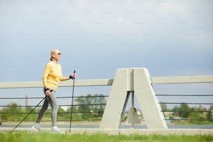 AGed sportswoman in pullover and leggins walking with trekking sticks in urban environment on summer day
