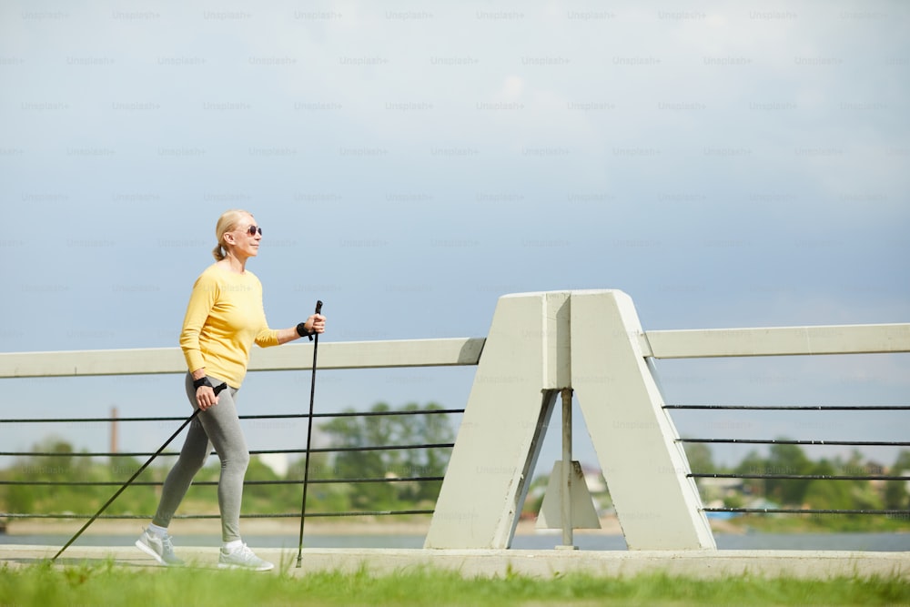 Deportista con jersey y leggins caminando con bastones de trekking en entorno urbano en día de verano