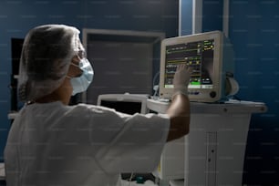 Asistente femenina joven con guantes, máscara y uniforme apuntando a la exhibición del equipo médico mientras toma el control de la condición corporal del paciente