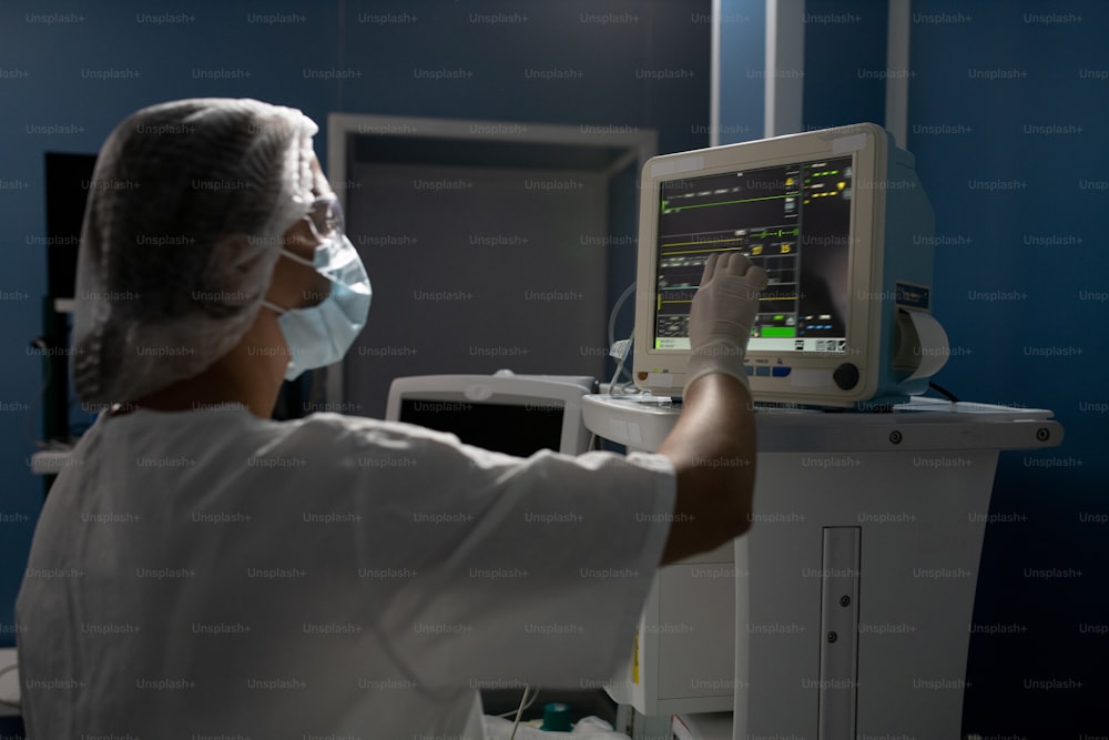 Jeune assistante en gants, masque et uniforme pointant l’étalage de l’équipement médical tout en prenant le contrôle de l’état corporel du patient