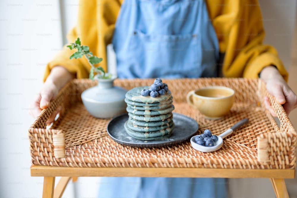 Frau in blauer Schürze mit Rattantablett mit blauen amerikanischen Pfannkuchen mit Blaubeere mit Karamellsouse, Tasse Kaffee und Vase mit Blume. Hausgemachtes leckeres Essen. Feier der Fastnacht.