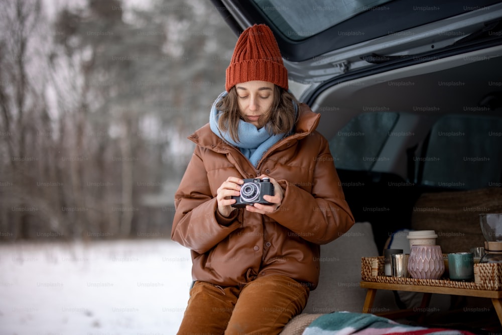 Un retrato de una mujer joven con una cámara vieja sentada en el maletero del automóvil, mirando su cámara en las manos, viajando en automóvil durante las vacaciones de invierno. Foto de alta calidad