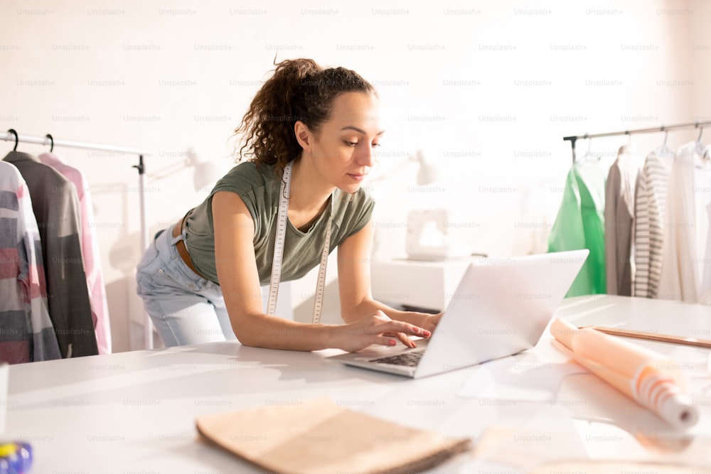 Joven diseñadora de moda inclinada sobre la mesa mientras se concentra en la búsqueda de datos en línea