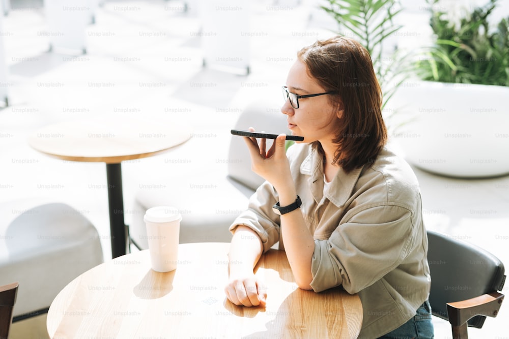 Jeune adolescente brune étudiante à lunettes utilisant un téléphone portable a envoyé un message vocal assis dans un café moderne vert