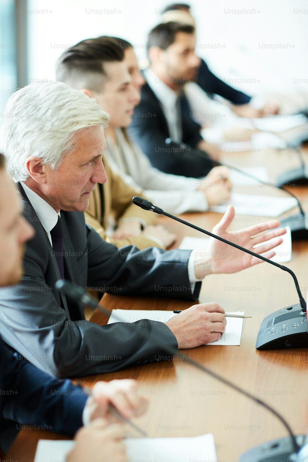 Mature grey-haired delegate speaking in microphone while commenting speech of one of participants of summit