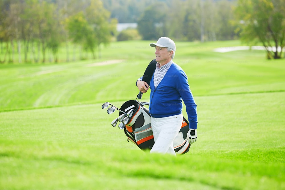 Homem sênior em casualwear andando para o jogo de golfe anf carregando tacos no saco
