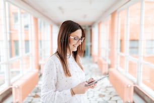 Portrait of young smiling Caucasian female teacher with eyeglasses using tablet while standing in the hall. Side view.