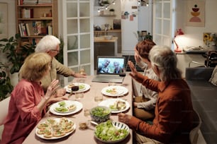 Grupo de amigos mayores agitando las manos mientras saludan a la pareja de ancianos en la pantalla de la computadora portátil durante la comunicación en video chat por mesa