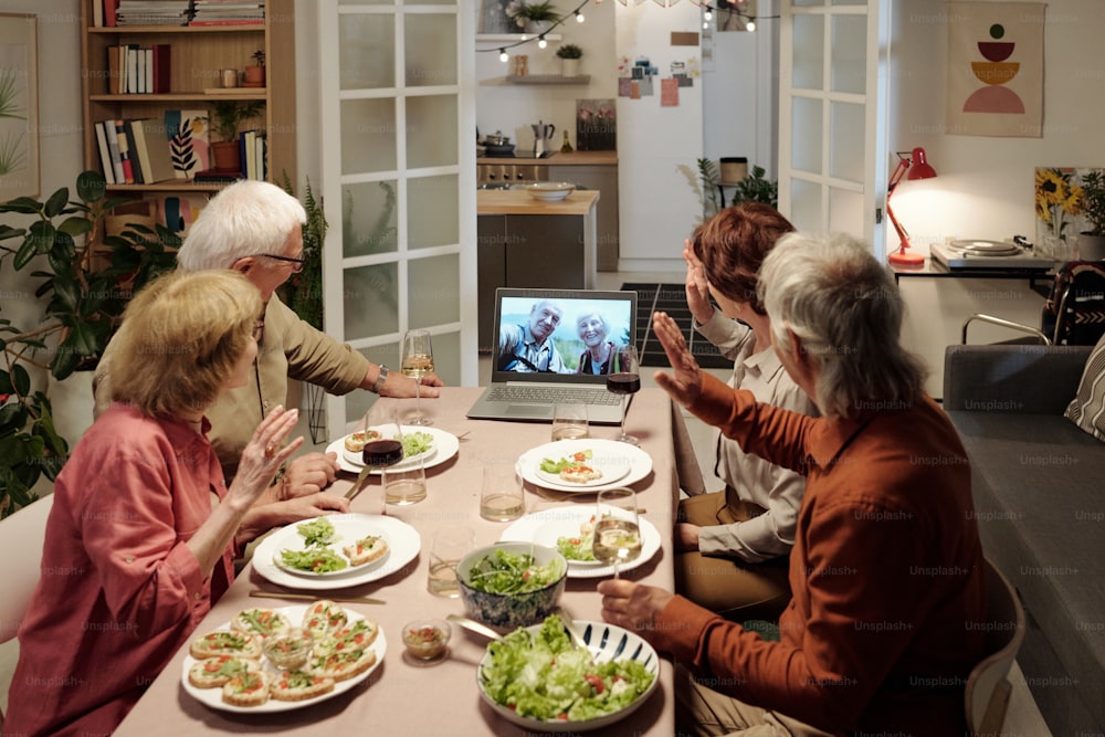 Group of senior friends waving hands while greeting aged couple on screen of laptop during communication in video chat by table