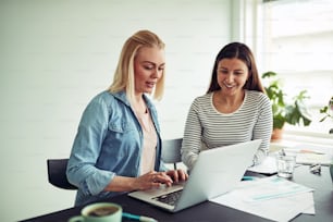 Two smiling young businesswomen sitting at a desk in an office talking together and working with a laptop