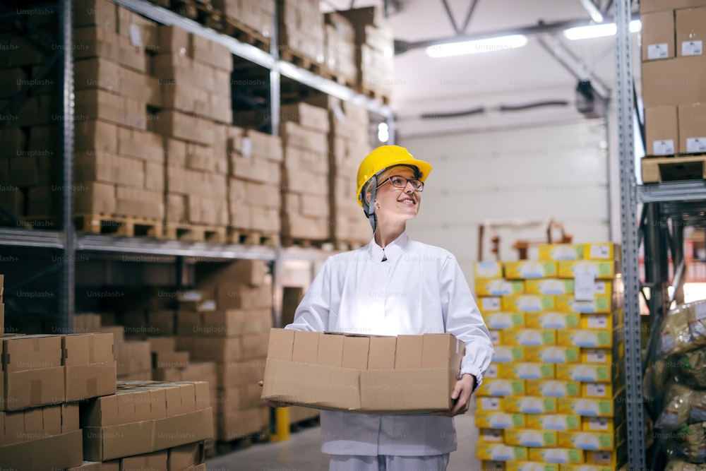 Smiling female worker carrying boxes in warehouse. Protective helmet on head, all around boxes on shelves.