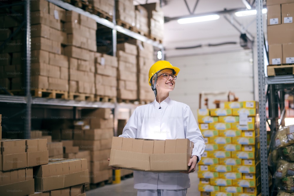Travailleuse souriante portant des boîtes dans l’entrepôt. Casque de protection sur la tête, tout autour des boîtes sur les étagères.