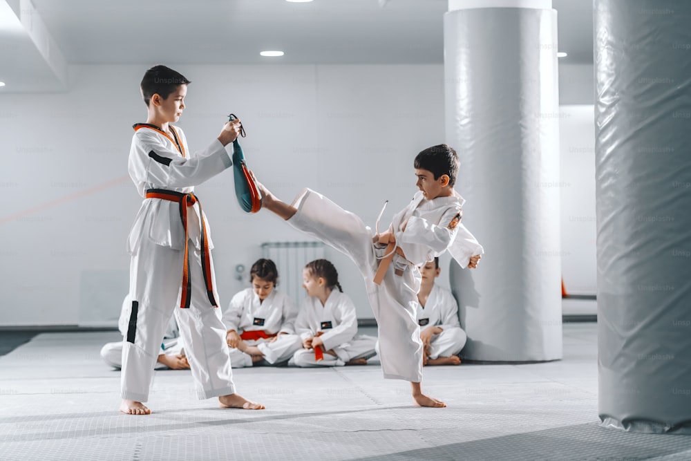 Dos jóvenes caucásicos en doboks entrenando taekwondo en el gimnasio. Un niño pateando mientras otro sosteniendo el objetivo de patada. En el fondo, su amigo sentado con las piernas cruzadas y observándolos.