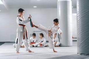 Zwei junge kaukasische Jungen in Doboks beim Taekwondo-Training im Fitnessstudio. Ein Junge tritt, während der andere das Ziel hält. Im Hintergrund sitzt ihr Freund mit gekreuzten Beinen und beobachtet sie.