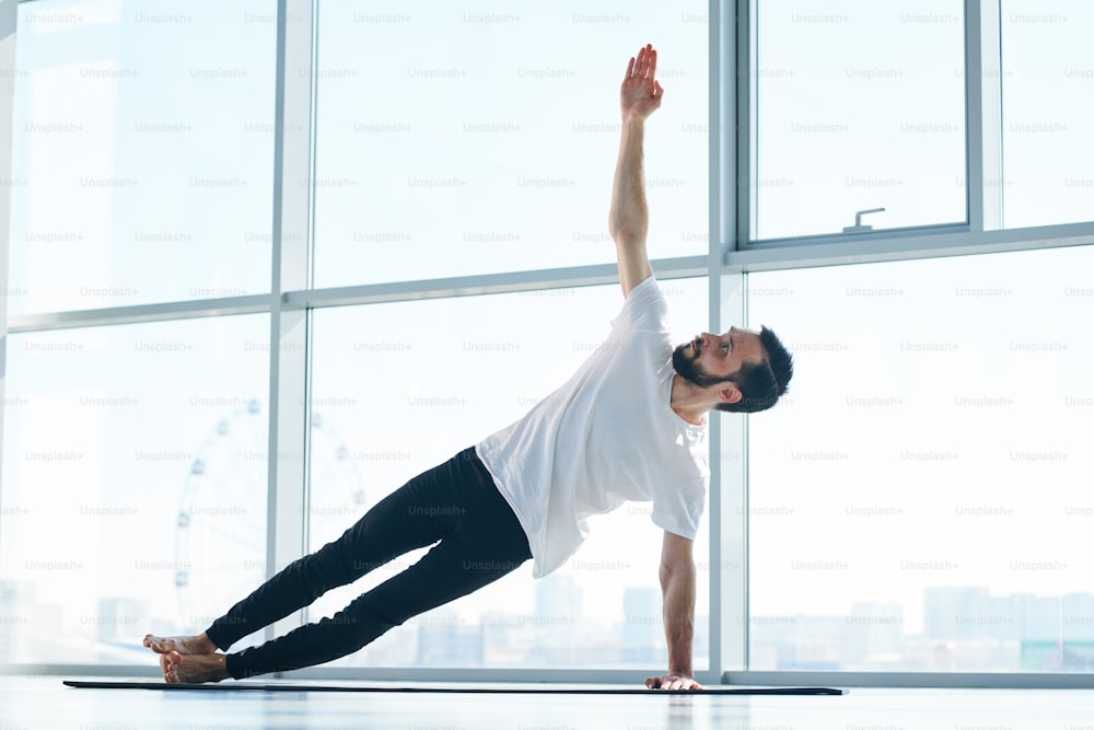 Deportista activo con camiseta blanca y leggins negros levantando el brazo estirado sobre la cabeza durante uno de los ejercicios de yoga en el suelo