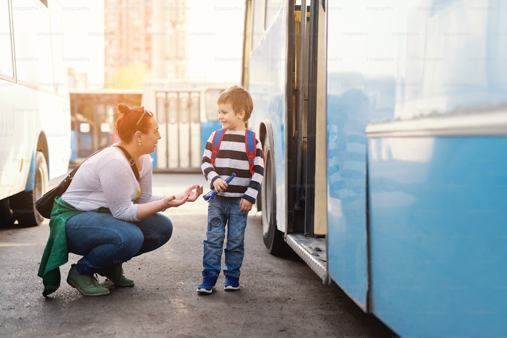 Madre diciendo hablando con su hijo mientras está agachada frente al autobús. Niño yendo a la escuela.