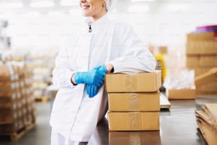 Young joyful female worker is leaned against the stack of boxes in a factory storage room.