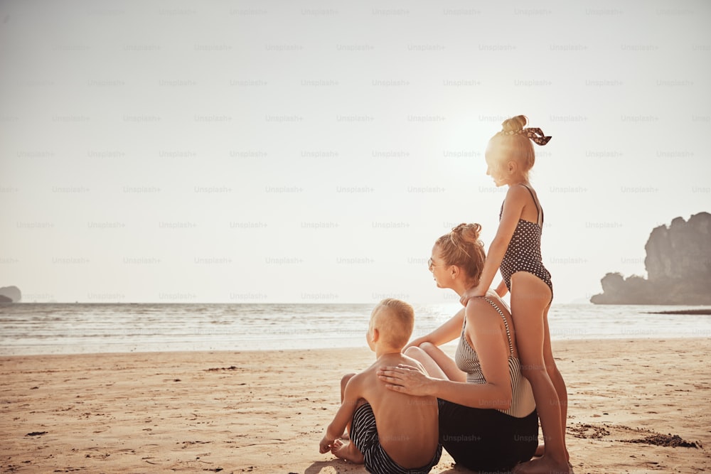 Mãe sorridente e seus dois filhos olhando para o oceano enquanto sentados juntos em uma praia de areia durante as férias de verão