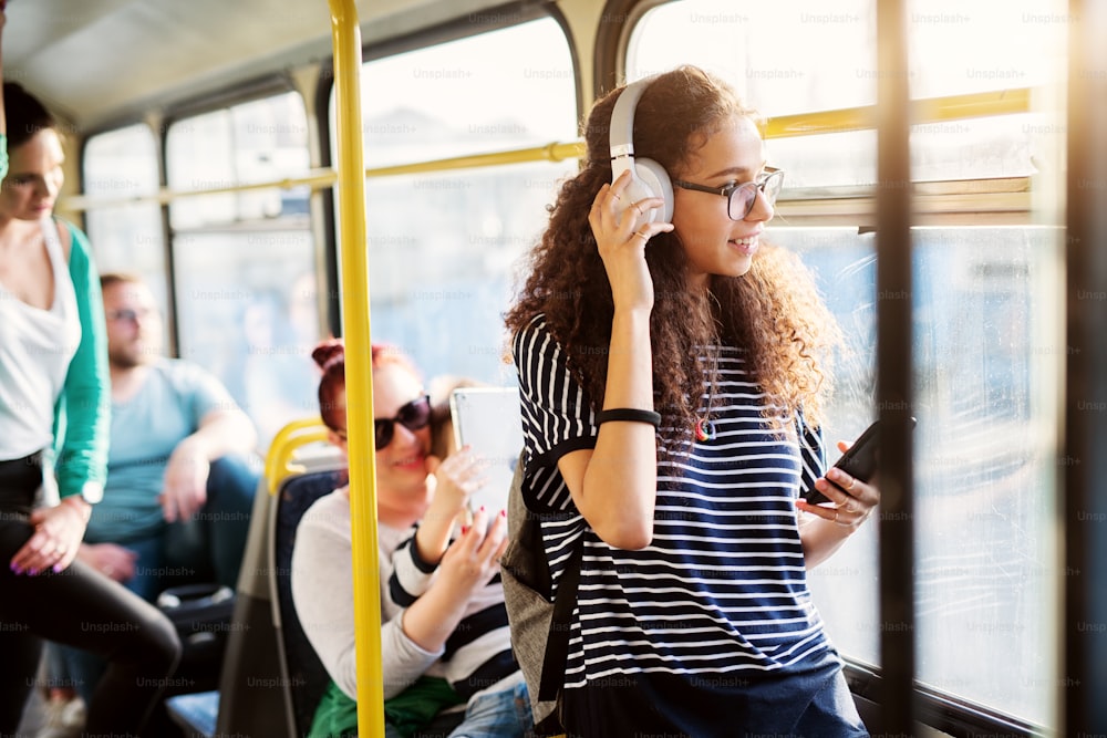 Giovane bella donna con i capelli ricci sta regolando le sue cuffie ascoltando musica e usando il telefono mentre viaggia in autobus e guarda attraverso il finestrino.