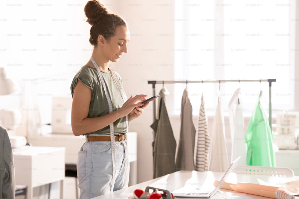 Cheerful self-employed fashion designer scrolling in smartphone during work in design studio