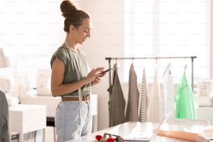 Cheerful self-employed fashion designer scrolling in smartphone during work in design studio