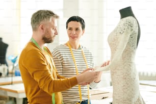 두 명의 전문 재단사가 스튜디오에서 더미 옆에 서 있는 동안 레이스 드레스의 새 모델에 대한 세부 사항에 대해 상담합니다.