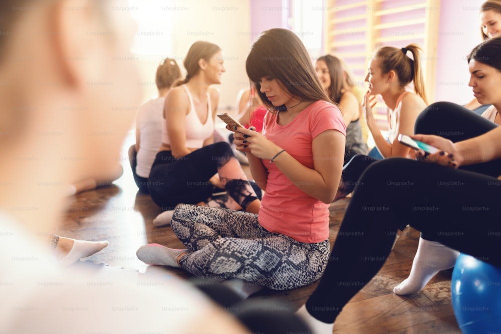 Group beautiful fit of sporty women sitting in gym and using smart phones and resting after exercises. Selective focus on brunette in t-shirt.