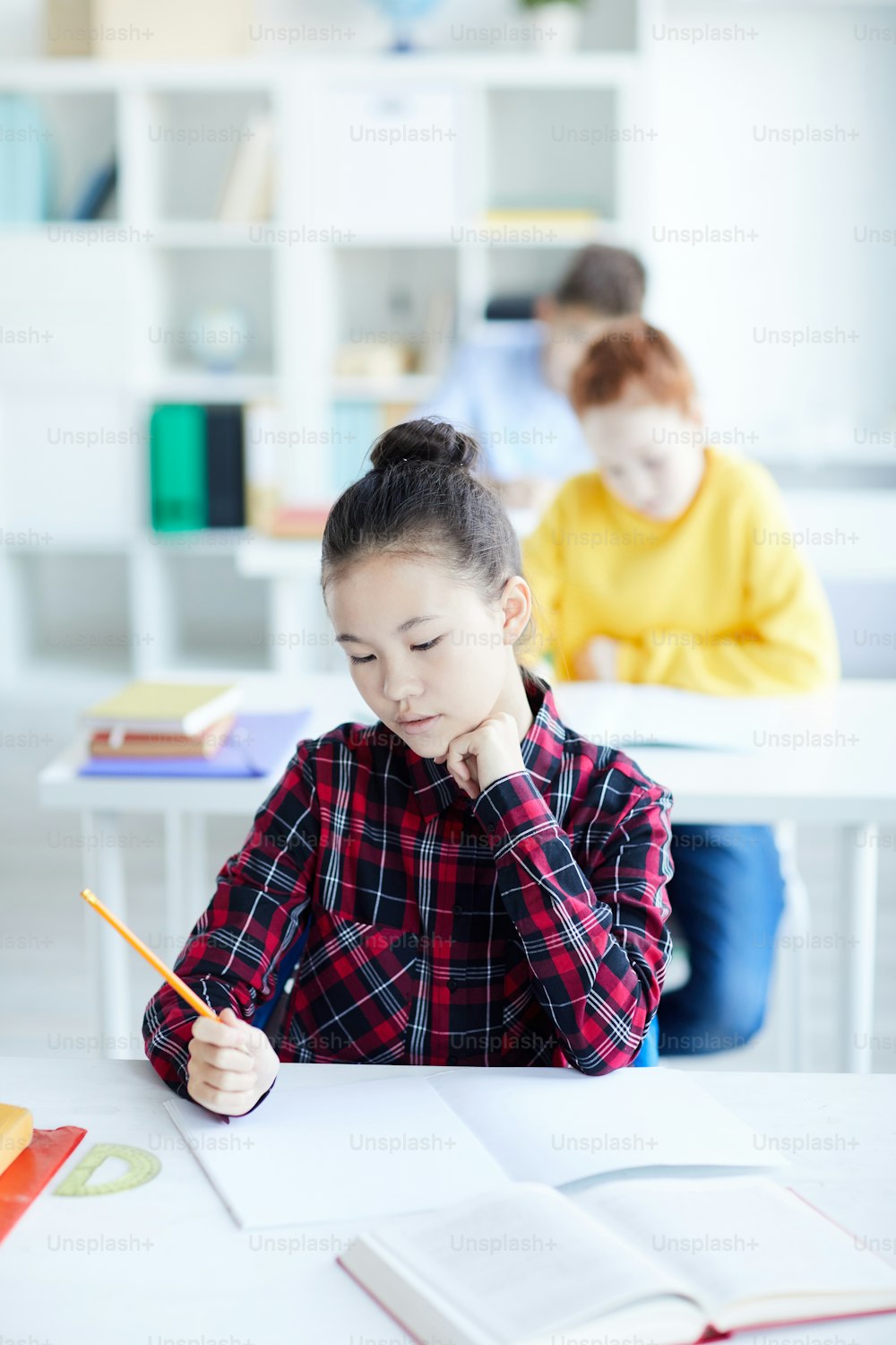 Pensive little girl with crayon sitting by desk in classroom and looking at blank page of copybook while drawing