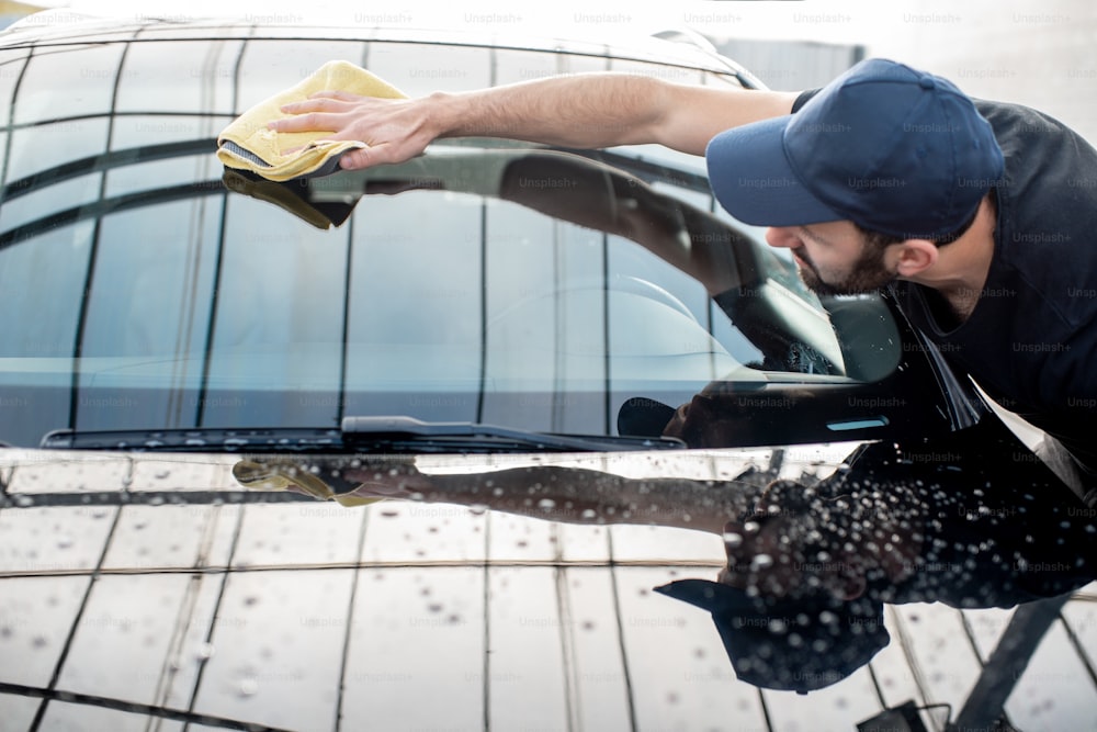 Professional washer in t-shirt and cap wiping windshield with yellow microfiber at the open air car wash