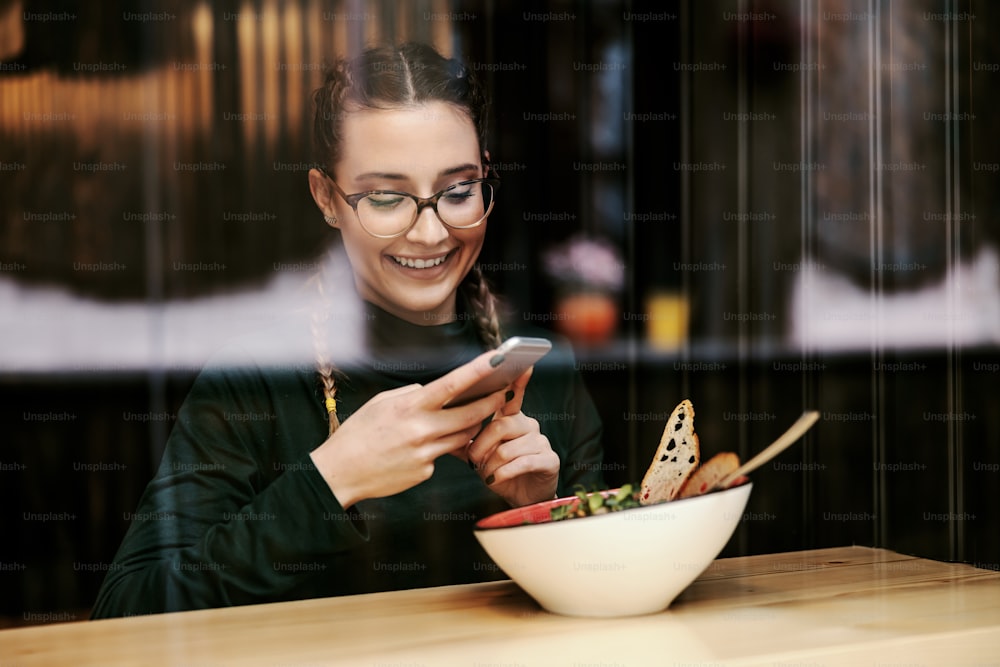 Jovem nerd sentada no restaurante e tirando foto de seu delicioso almoço saudável. Ela vai postá-lo nas redes sociais. Foto tirada de fora.