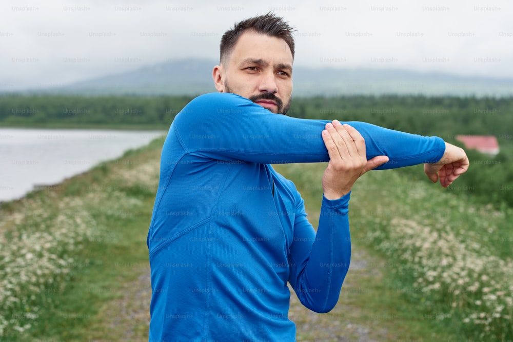 Mid age sportsman exercising outdoor, warming up arm muscules, in summer, on gloomy day, standing at the road with scenic view, doing warm up stretching, wearing blue jacket, he has a beard