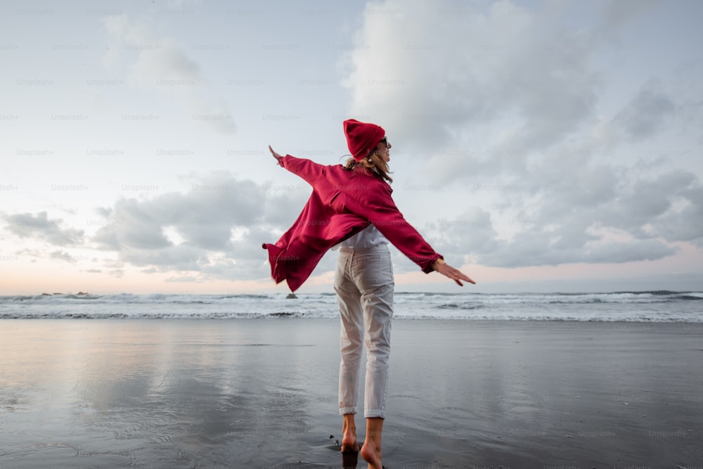 빨간 셔츠와 모자를 쓴 평온한 여자가 황혼에 해변을 걷고 있는 라이프스타일 초상화. 웰빙, 행복 및 삶의 즐거움 개념