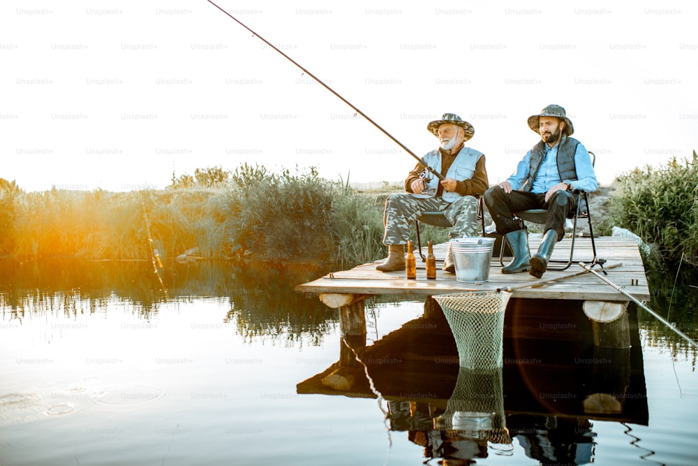 Grandfather with adult son fishing together on the wooden pier during the morning light. View from the side of the lake