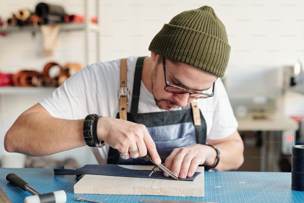 Giovane concentrato in cappello hipster che usa calibri nella lavorazione della pelle mentre misura il bordo del pezzo di pelle