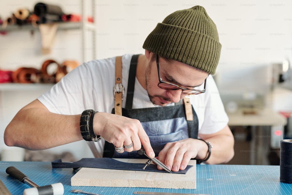 Konzentrierter junger Mann mit Hipster-Hut, der Bremssättel in der Lederverarbeitung benutzt, während er die Kante des Lederstücks misst