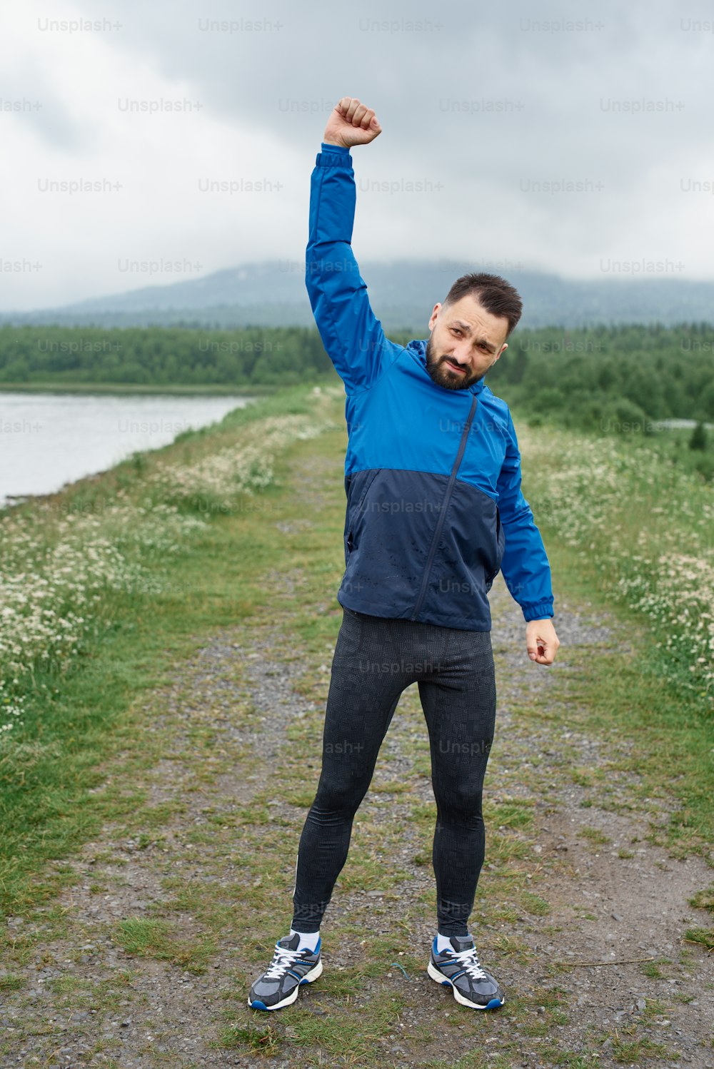Mid age sportsman exercising outdoor in summer, on gloomy day, standing at the road with scenic view, doing warm up stretching, wearing blue jacket, he has a beard