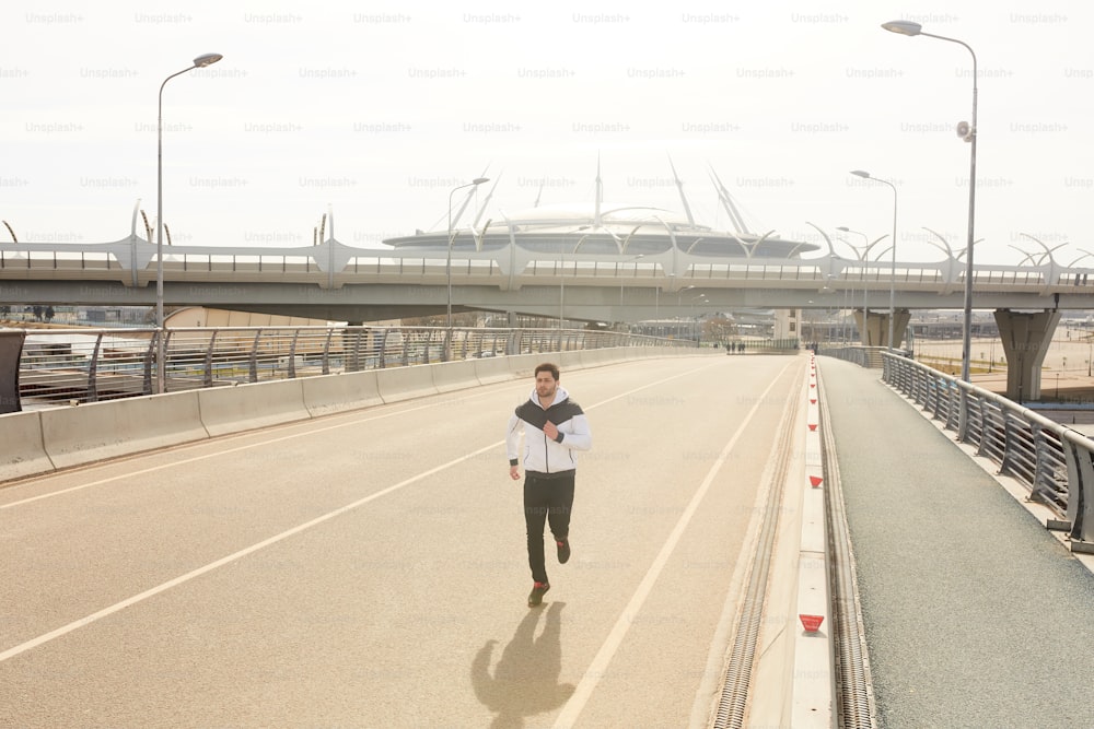 Young male runner jogging along racetrack along border during morning workout in urban environment