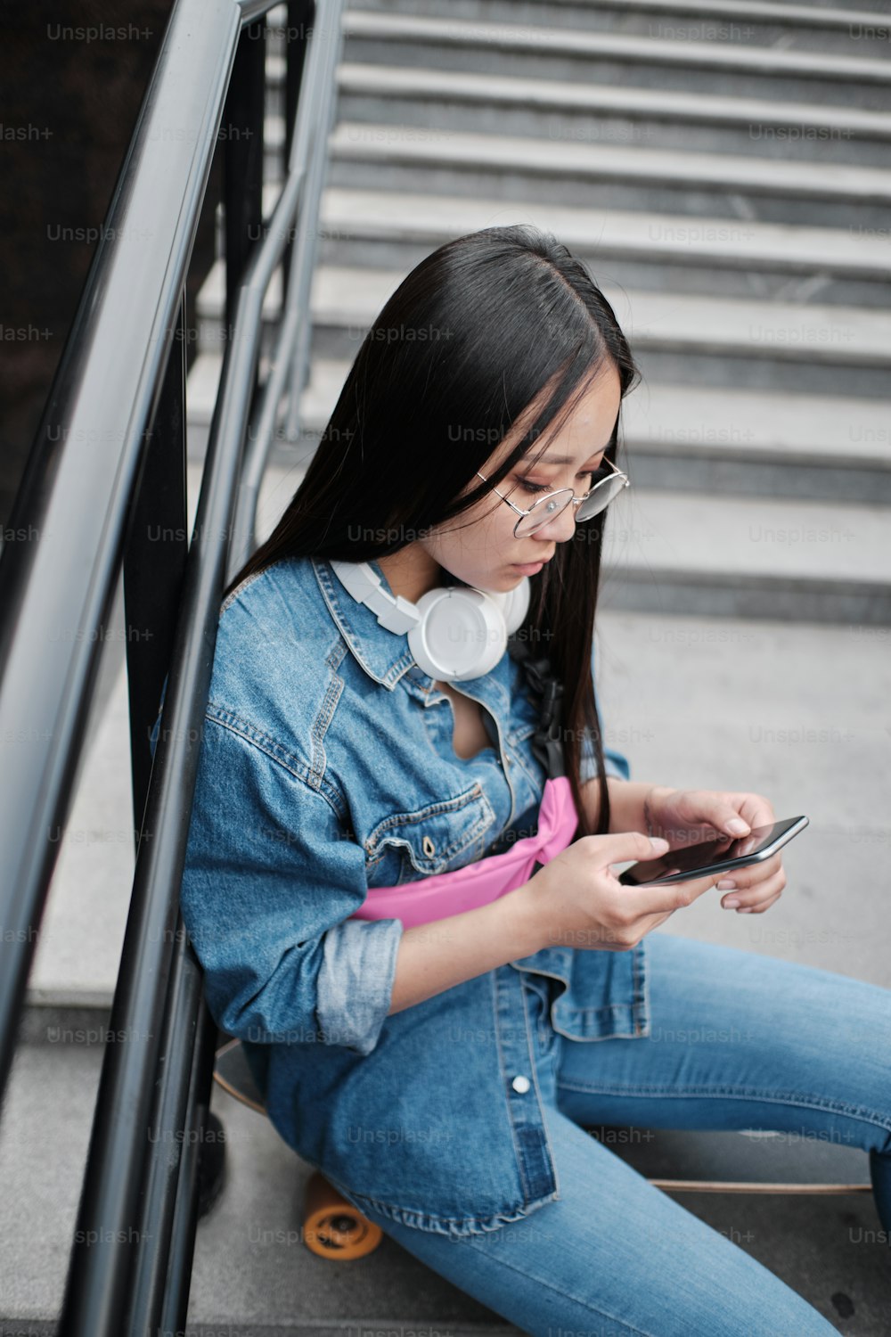 Close up of young asian skater girl sitting on her longboard on the stairs, using smart phone, wearing blue jeans, and pink belt bag, wearing round glasses and white headphones, looking away