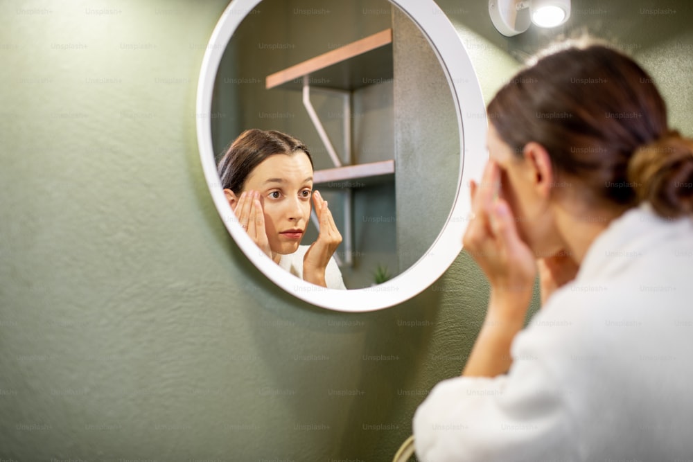 Woman in bathrobe making facial massage looking into the round mirror in the bathroom