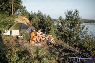 Coppia giovane e allegra che fa un picnic al campeggio mentre viaggia nella foresta vicino al lago durante il tramonto