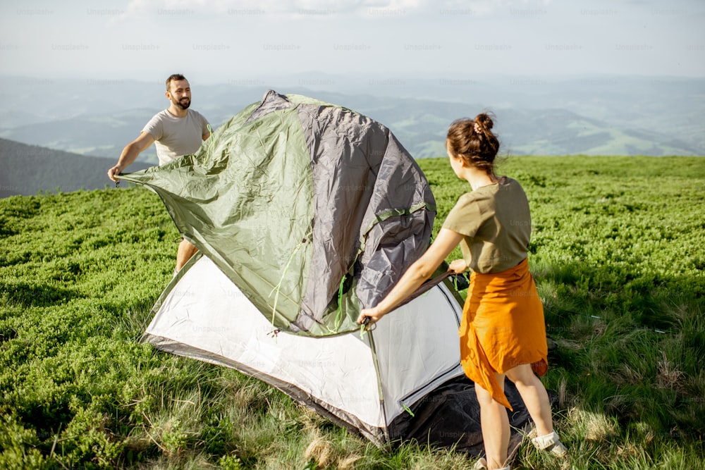 Young couple setting up the tent on the green meadow, traveling high in the mountains during the sunset